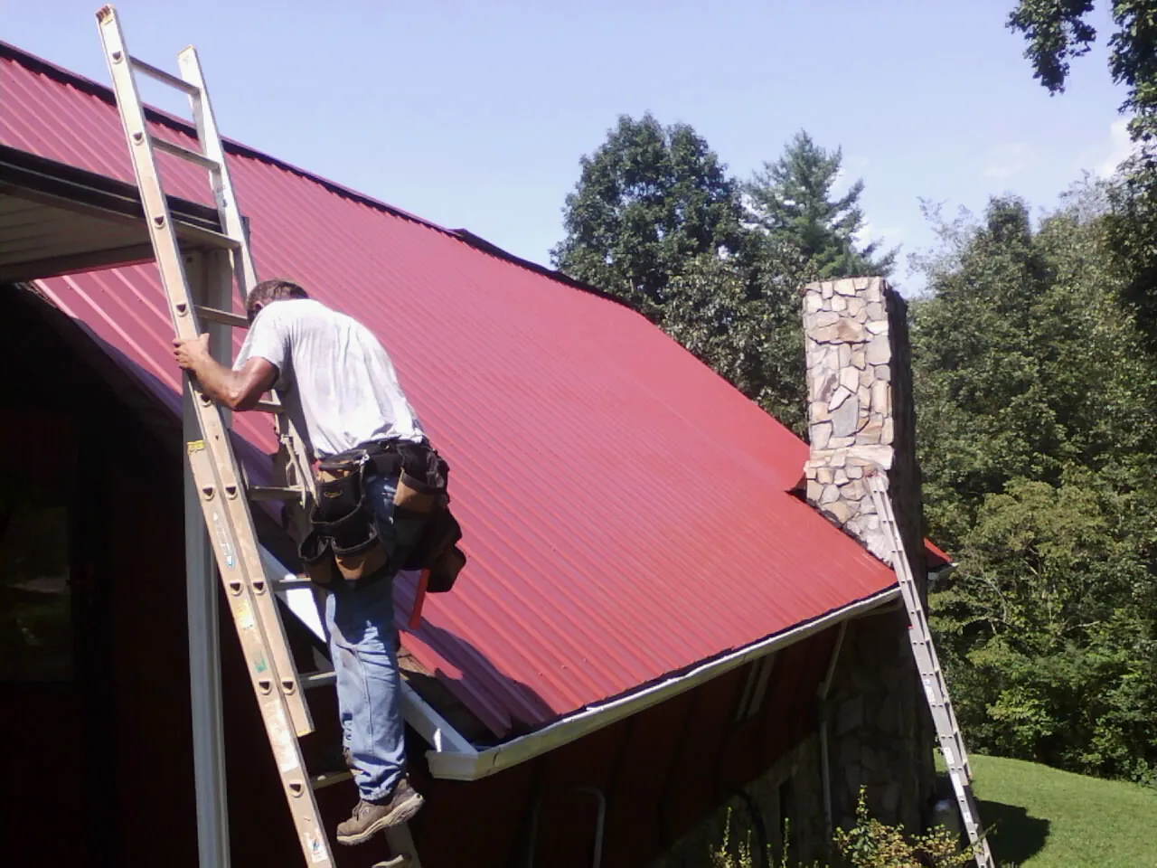 Roofer on a ladder beside a red metal roof and stone chimney — Soffit & Fascia Repair by Roofing Brothers in Mahnomen